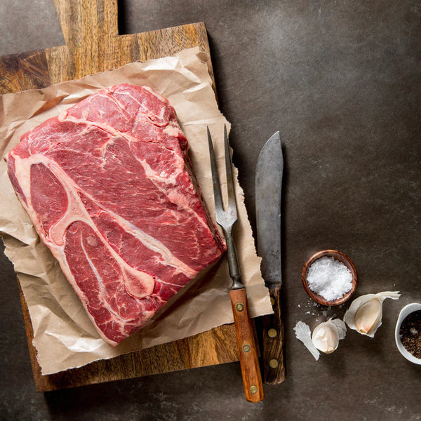 Raw steak on a wooden cutting board with a knife, salt, and garlic on a dark surface